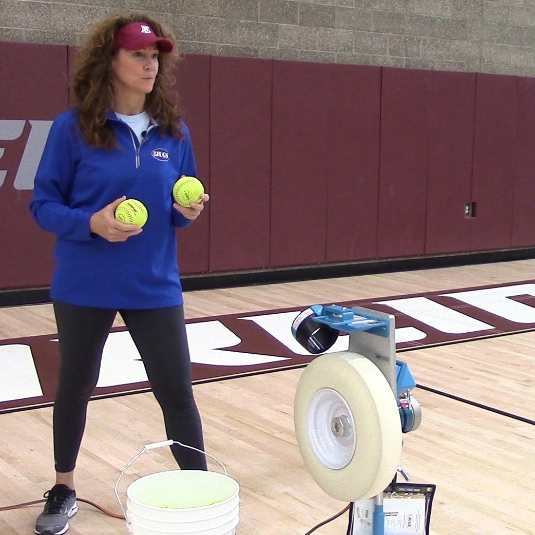 A coach holding two yellow JUGS LeatherLast softballs beside a JUGS Changeup pitching machine and a bucket of balls in a gym.