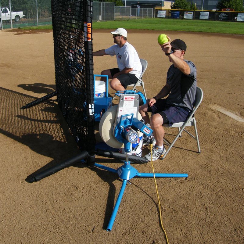 Two coaches seated behind a black protective net on a dirt infield, one feeding a yellow softball into a JUGS BP1 pitching machine.