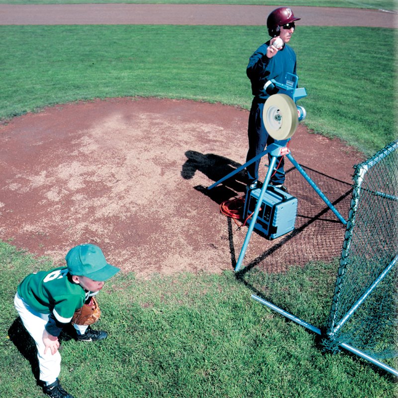 A JUGS pitching machine on tripod legs on a grass baseball field, with a young catcher crouching nearby and a player beside the machine.