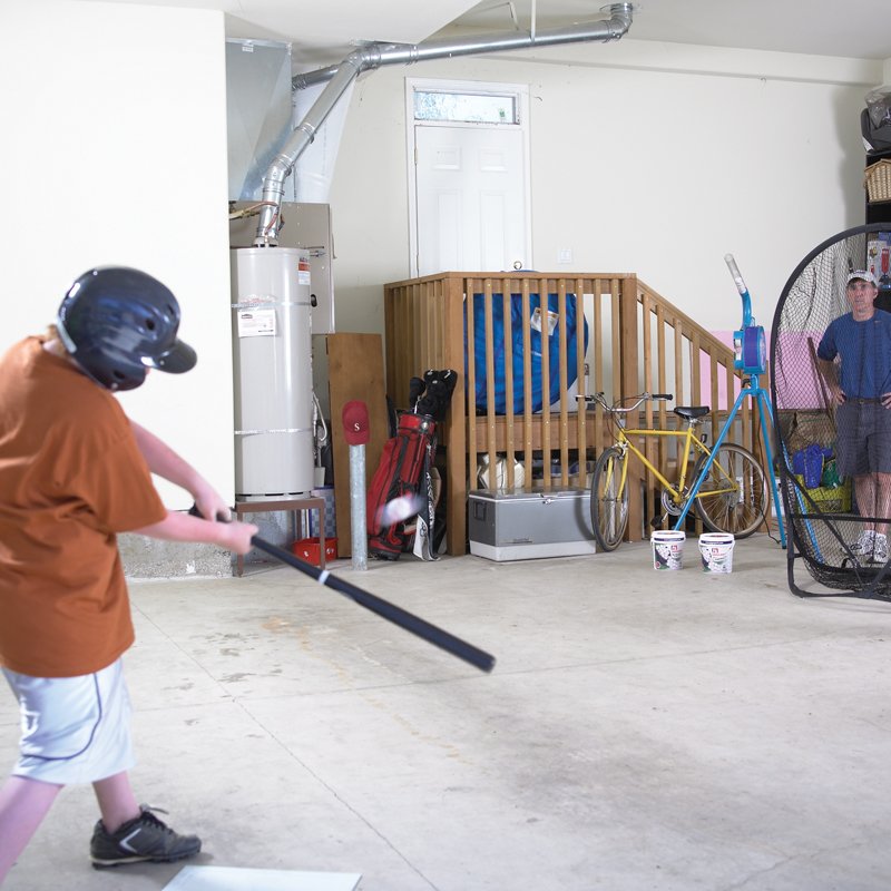 A boy in a batting helmet swinging indoors in a garage, practicing with small balls.
