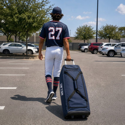 A baseball player in a navy jersey with the number 27 and white pants walks across a parking lot, pulling a navy rolling bat bag behind him by its extended handle.