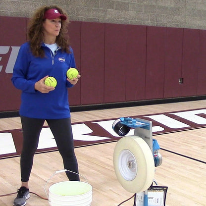 A coach holding two yellow JUGS LeatherLast softballs beside a JUGS Changeup pitching machine and a bucket of balls in a gym.