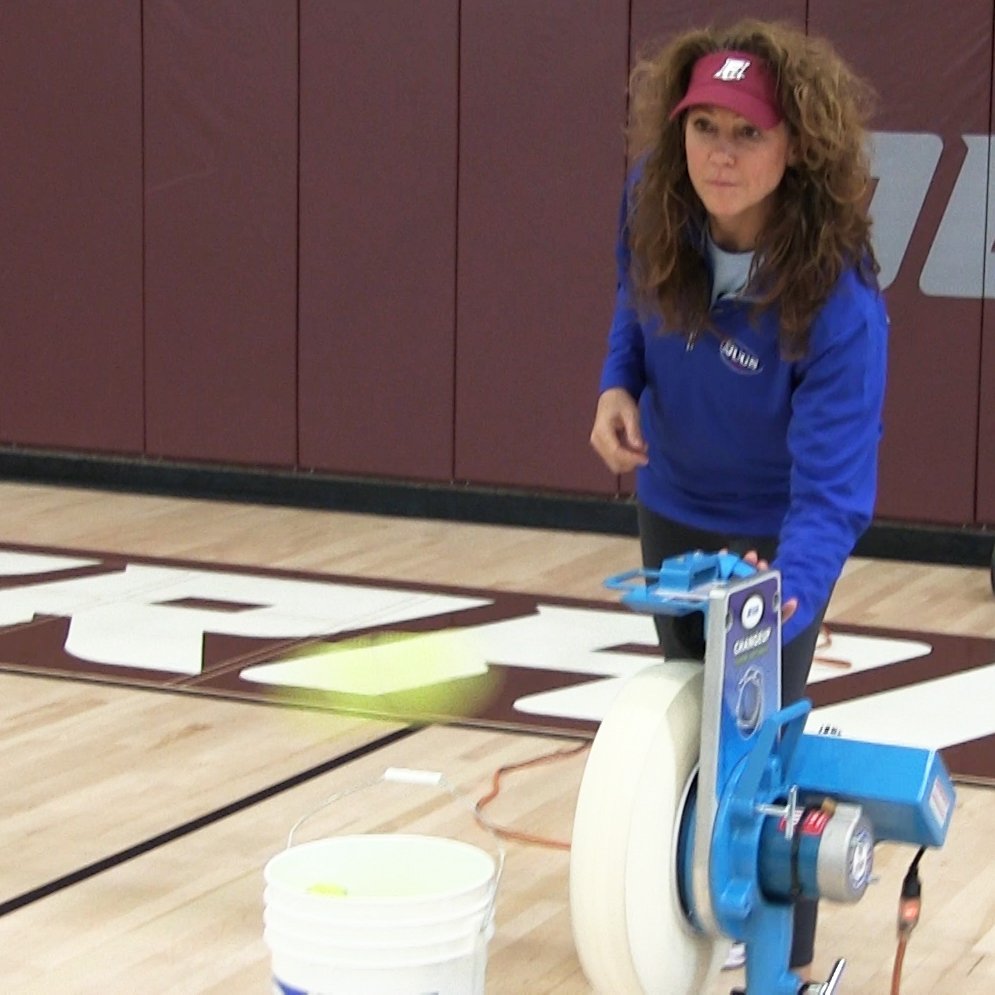 A coach feeding a yellow JUGS LeatherLast softball into a JUGS Changeup softball pitching machine in a gym, with a ball bucket beside the machine.