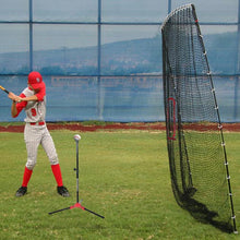 A young player in a red uniform hits off a batting tee into a tall black net standing on a grass field.