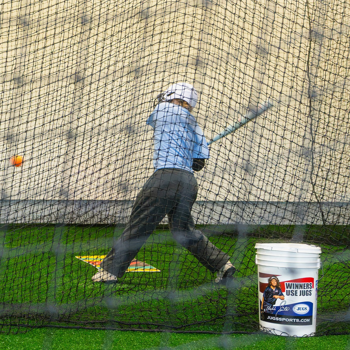 A young hitter swinging in a backyard JUGS batting cage net with a bucket of balls nearby.