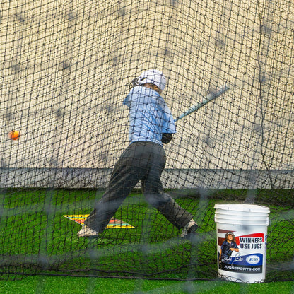 A young hitter swinging in a backyard JUGS batting cage net with a bucket of balls nearby.