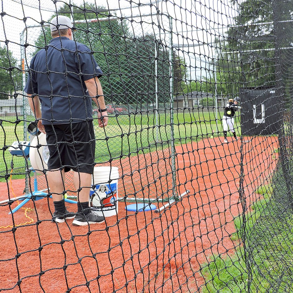 A coach running a JUGS pitching machine at the end of an outdoor JUGS batting cage with a backdrop hung at the far end.