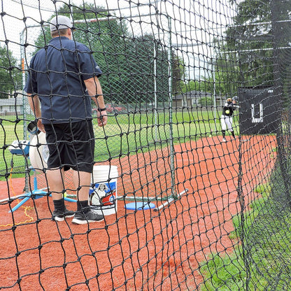 A coach running a JUGS pitching machine at the end of an outdoor JUGS batting cage with a backdrop hung at the far end.