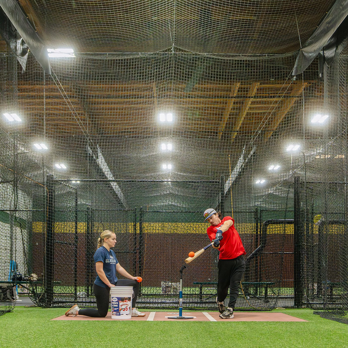Two softball players working a hitting station inside a JUGS batting cage net in a lit indoor facility.