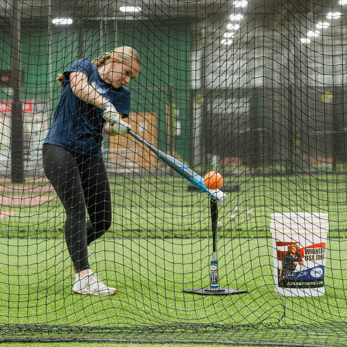 A softball player hitting off a tee inside a JUGS batting cage net in an indoor training facility.
