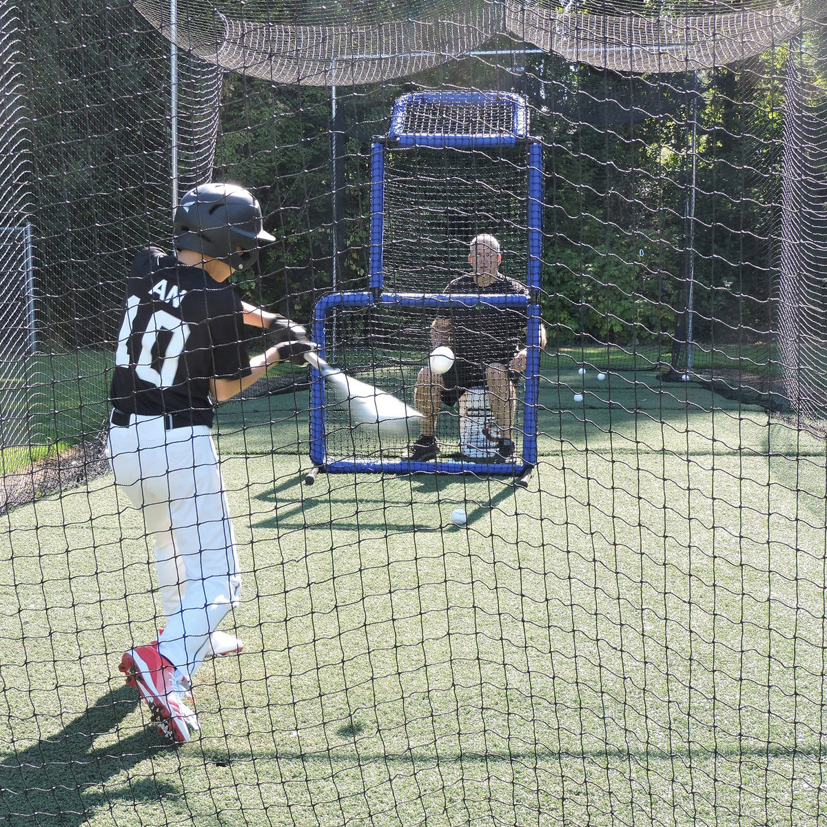 A young baseball player swinging in a JUGS batting cage net with a coach seated behind an L-screen.