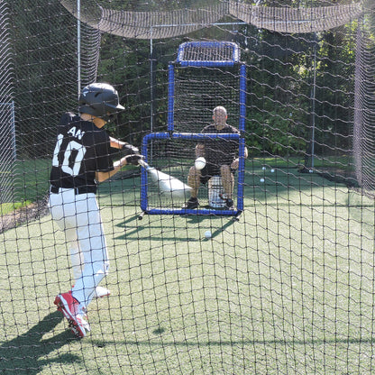 A young baseball player swinging in a JUGS batting cage net with a coach seated behind an L-screen.