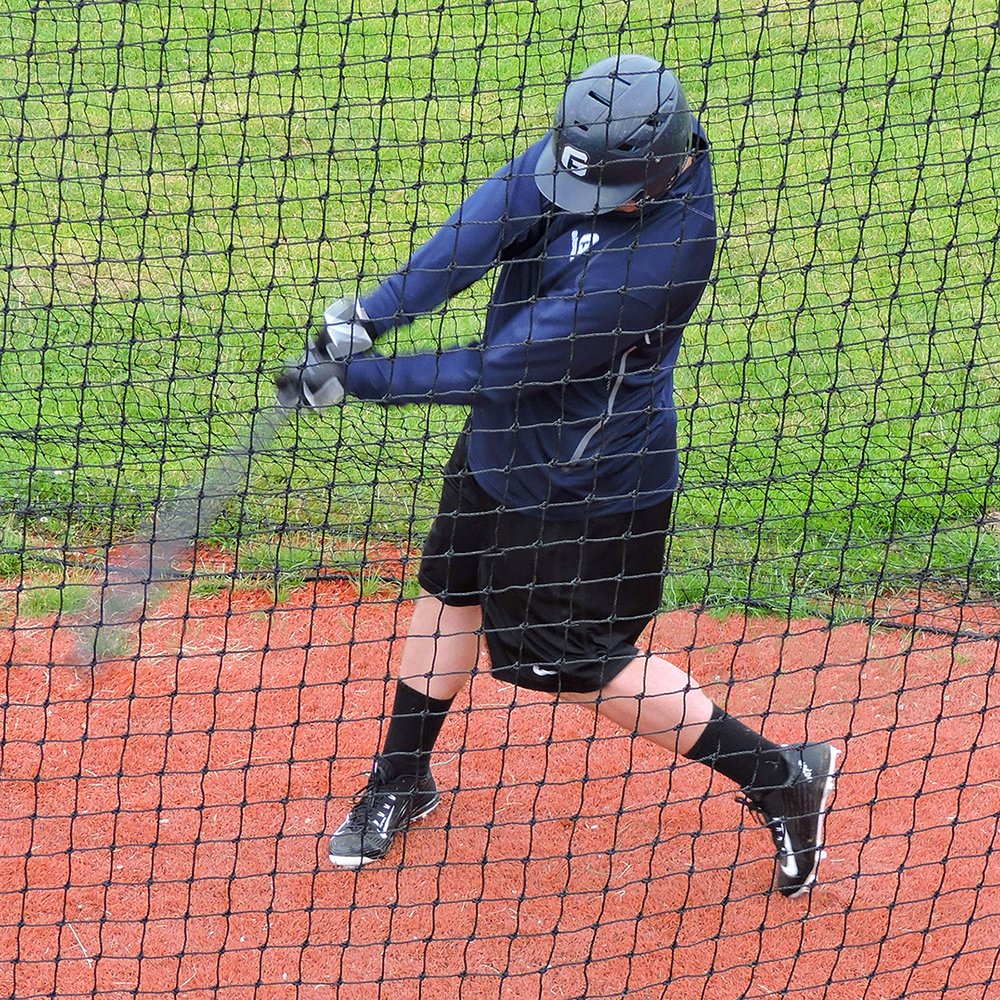 A baseball hitter in a batting helmet finishing a swing inside a JUGS batting cage net.