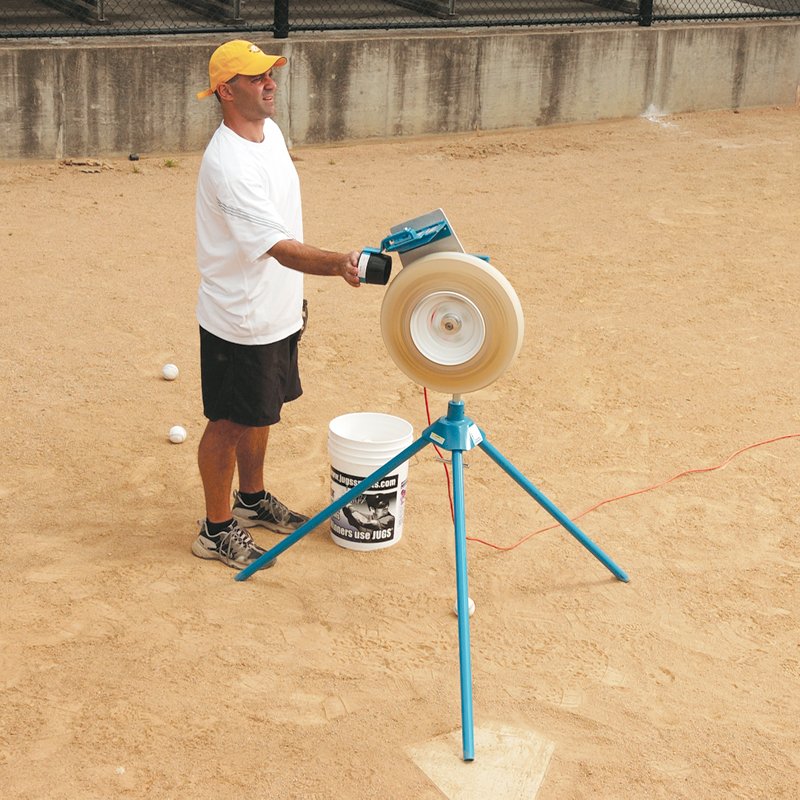 A coach in a white shirt and yellow cap loading a ball into a JUGS pitching machine on tripod legs on a dirt infield, with a white bucket behind him.