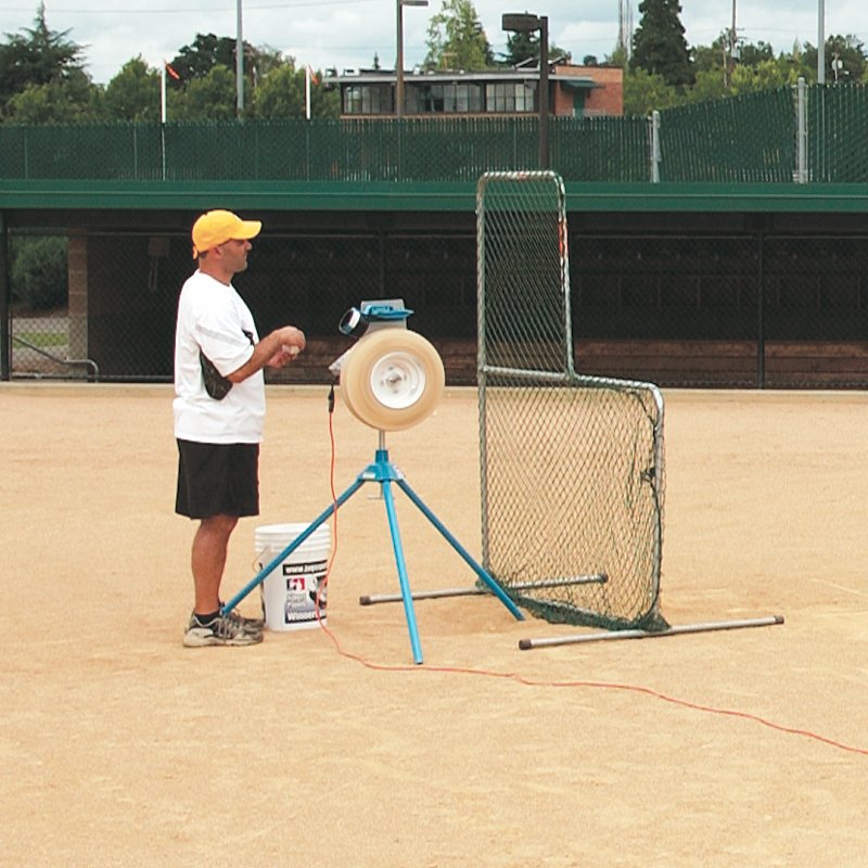 A coach in a yellow cap feeding a ball into a JUGS pitching machine on a dirt field, with an L-shaped protective screen and a bucket of balls nearby.