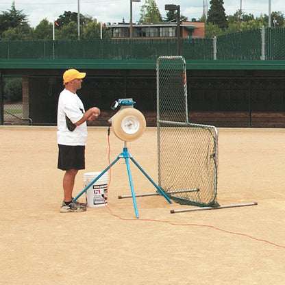 A coach in a yellow cap feeding a ball into a JUGS pitching machine on a dirt field, with an L-shaped protective screen and a bucket of balls nearby.