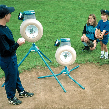 A coach holding a ball beside two JUGS pitching machines on a grass and dirt field while two young players kneel and watch.
