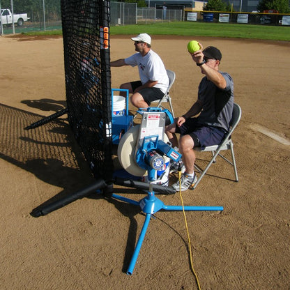 Two coaches seated behind a black protective net on a dirt infield, one feeding a yellow softball into a JUGS BP1 pitching machine.