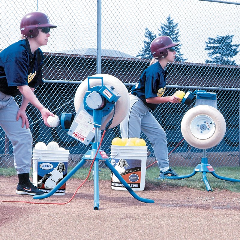 Two young players in batting helmets feeding softballs into two JUGS BP1 pitching machines set up side by side on a dirt field.