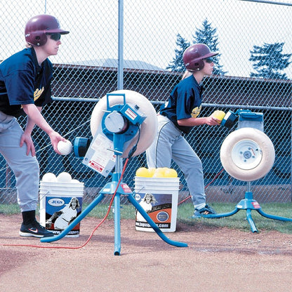 Two young players in batting helmets feeding softballs into two JUGS BP1 pitching machines set up side by side on a dirt field.