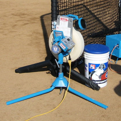 A JUGS BP1 pitching machine on tripod legs set up on a dirt infield next to a protective net, with a power cord and a bucket of balls beside it.