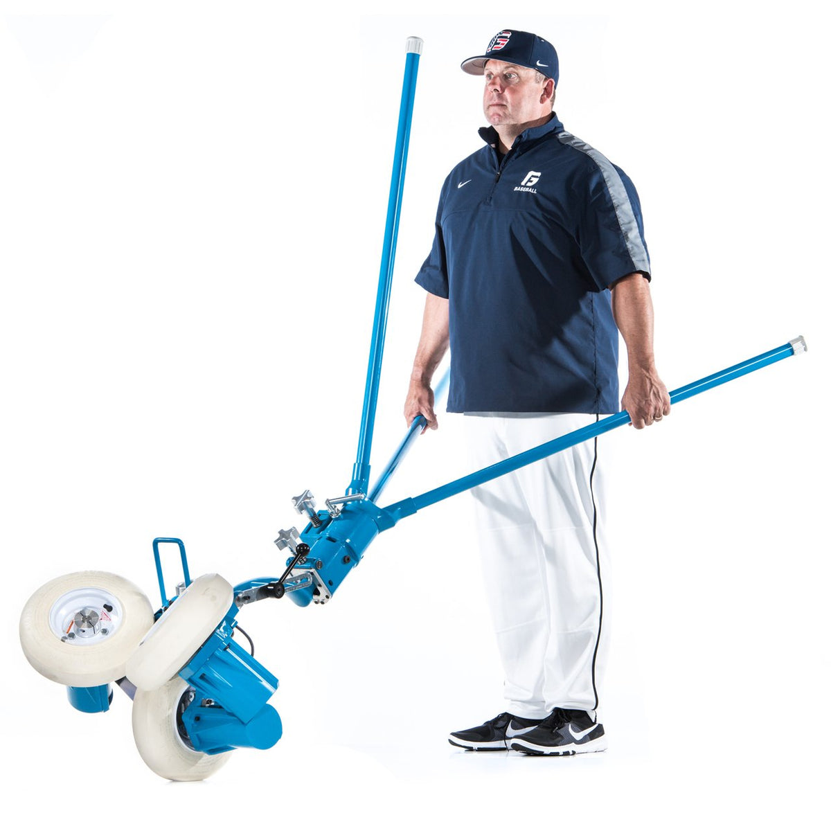 A man in a coaching uniform carrying a JUGS BP3 baseball pitching machine by its leg handles, photographed on a white background.