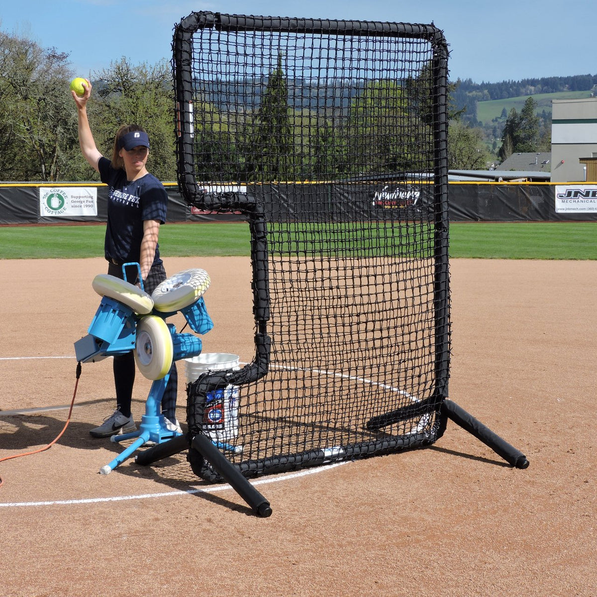 A JUGS BP3 softball pitching machine set up behind a C-shaped protective screen, with a player holding a softball up to feed it.
