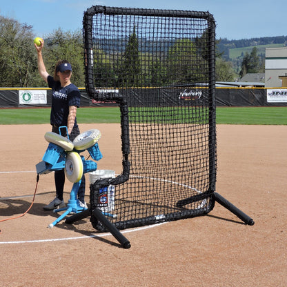 A JUGS BP3 softball pitching machine set up behind a C-shaped protective screen, with a player holding a softball up to feed it.