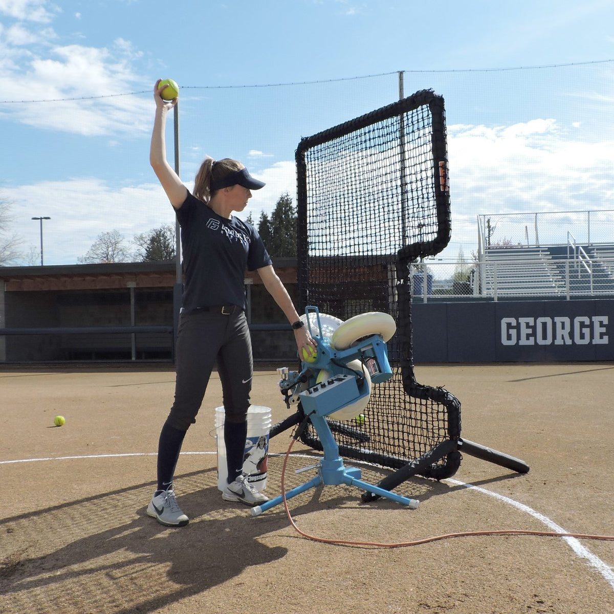 A softball player feeding a JUGS BP3 softball machine behind a C-shaped protective screen on a softball field.