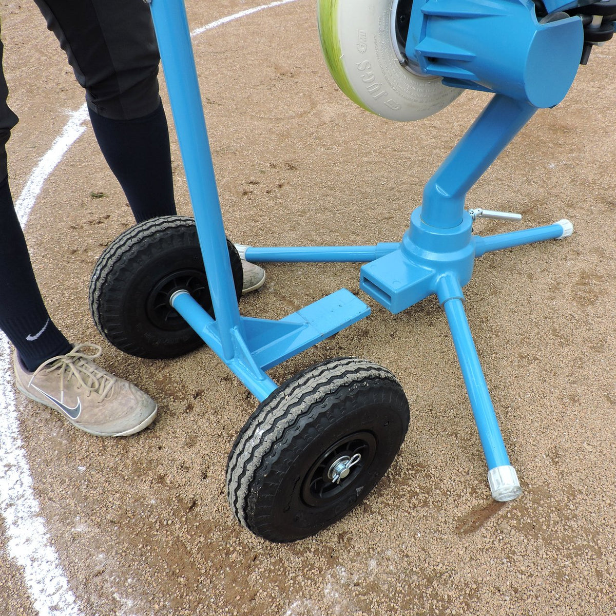 Close-up of the JUGS BP3 softball transport cart base with two black pneumatic wheels and light blue legs on infield dirt.