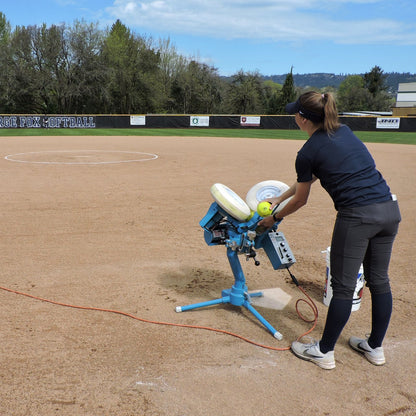 A coach loading a softball into a JUGS BP3 softball pitching machine set up on a softball diamond infield.