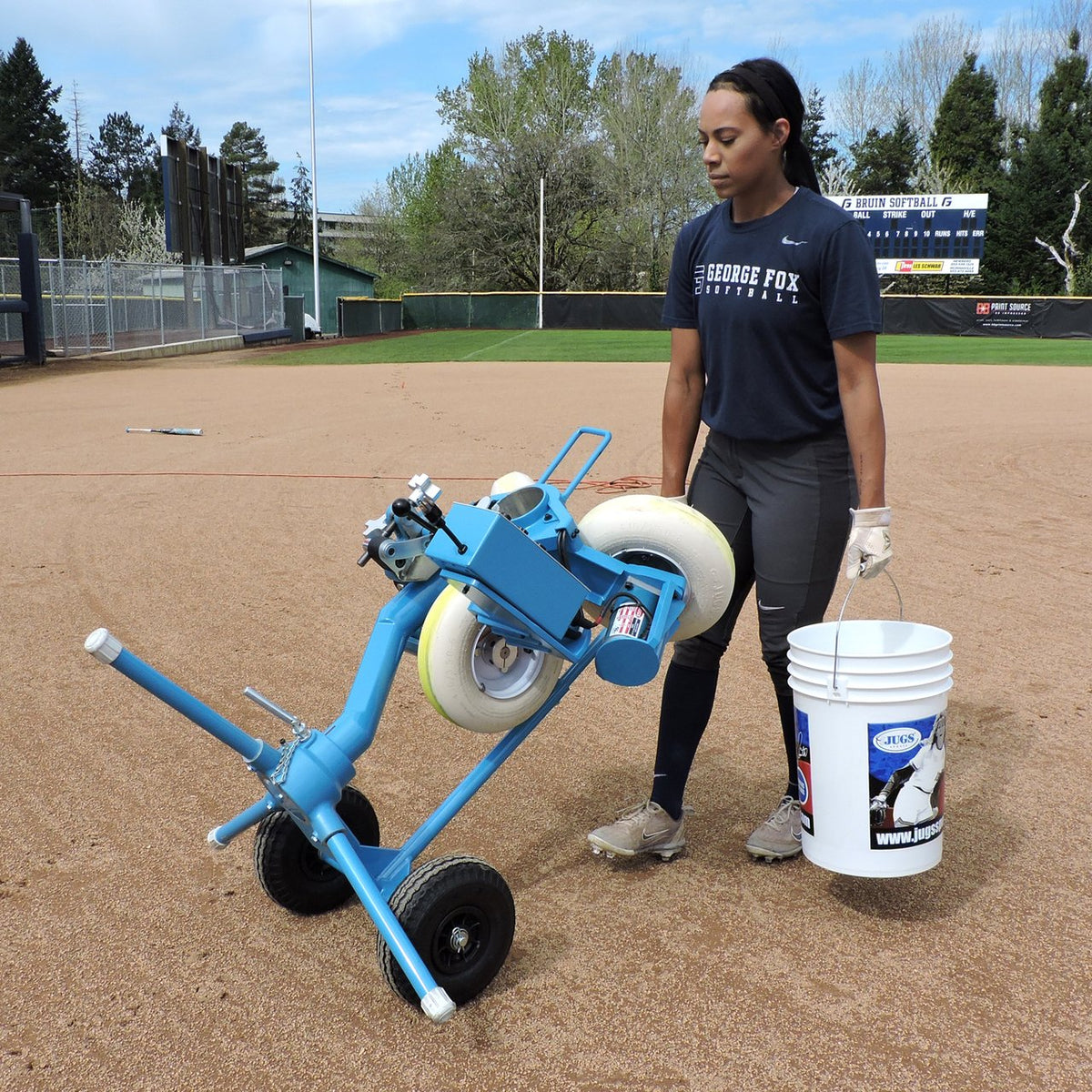 A softball player tilting a JUGS BP3 softball machine on its transport cart to roll it across the infield dirt, with a bucket of softballs beside her.