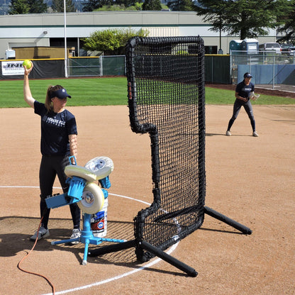 A coach feeding a JUGS BP3 softball pitching machine on a softball infield, with a C-shaped protective screen in front and a fielder behind.