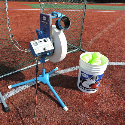A JUGS Changeup Super Softball pitching machine on tripod legs on a red clay infield beside a net, with a bucket of yellow softballs next to it.