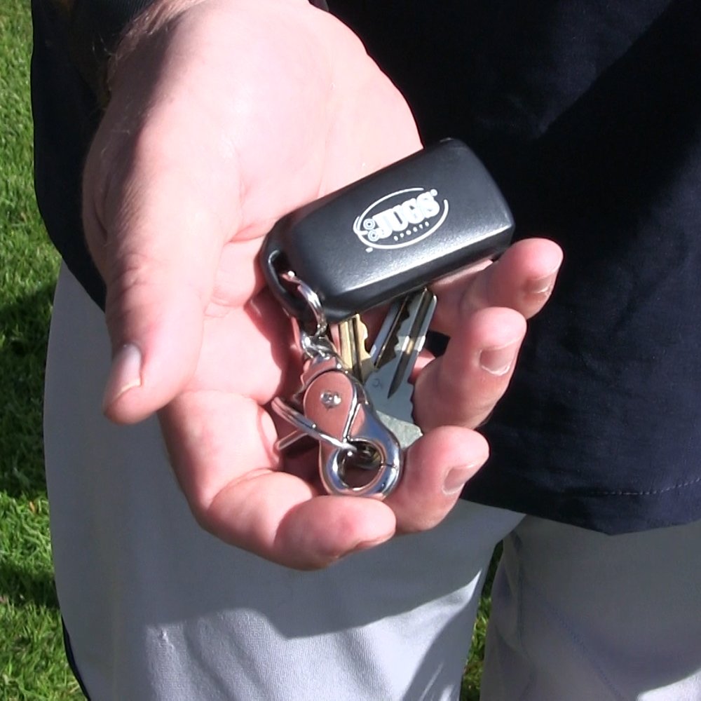 A hand holding a small black JUGS handheld remote control on a key ring, outdoors.