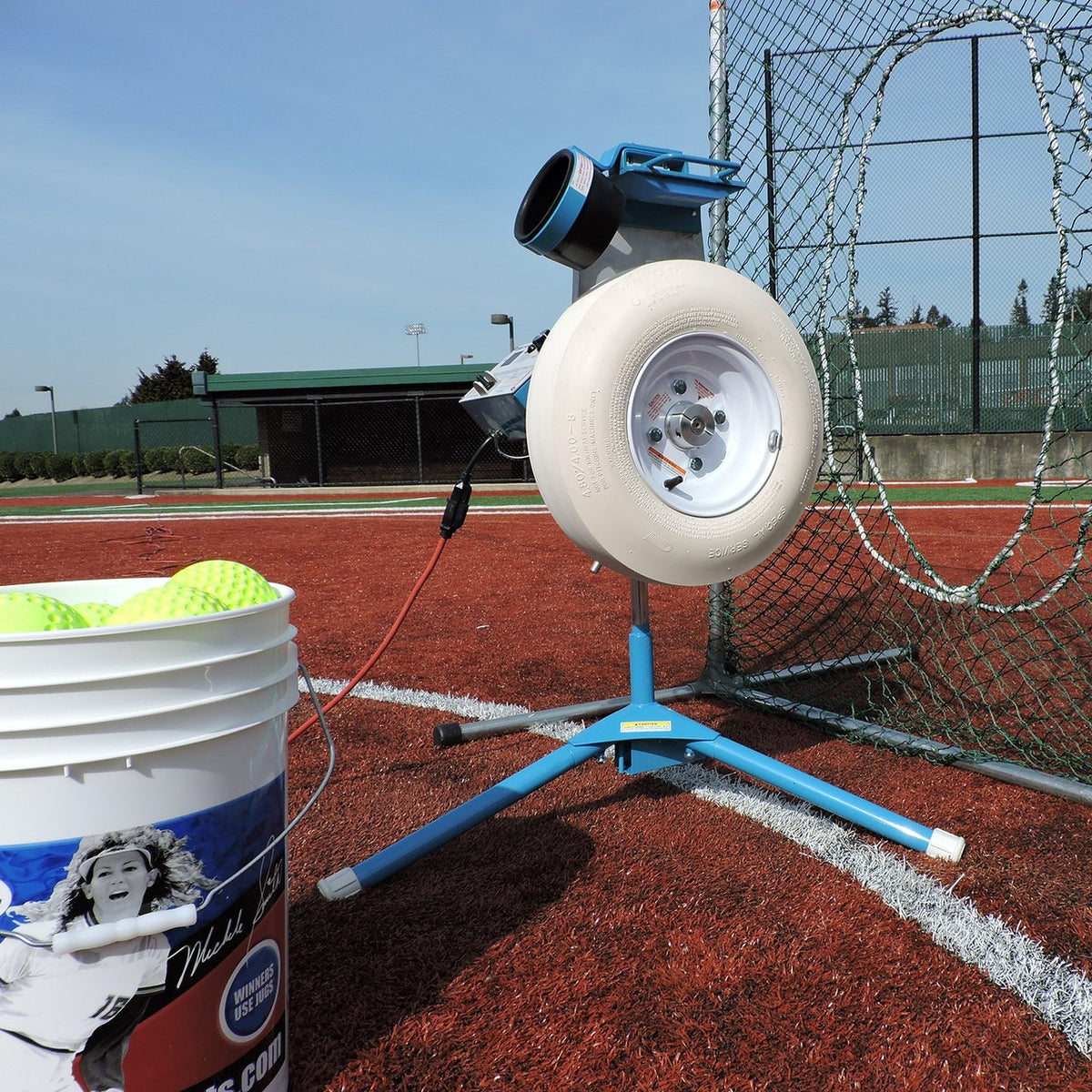 A side view of the JUGS Changeup Super Softball pitching machine on tripod legs on a clay infield, with a bucket of yellow softballs in the foreground.