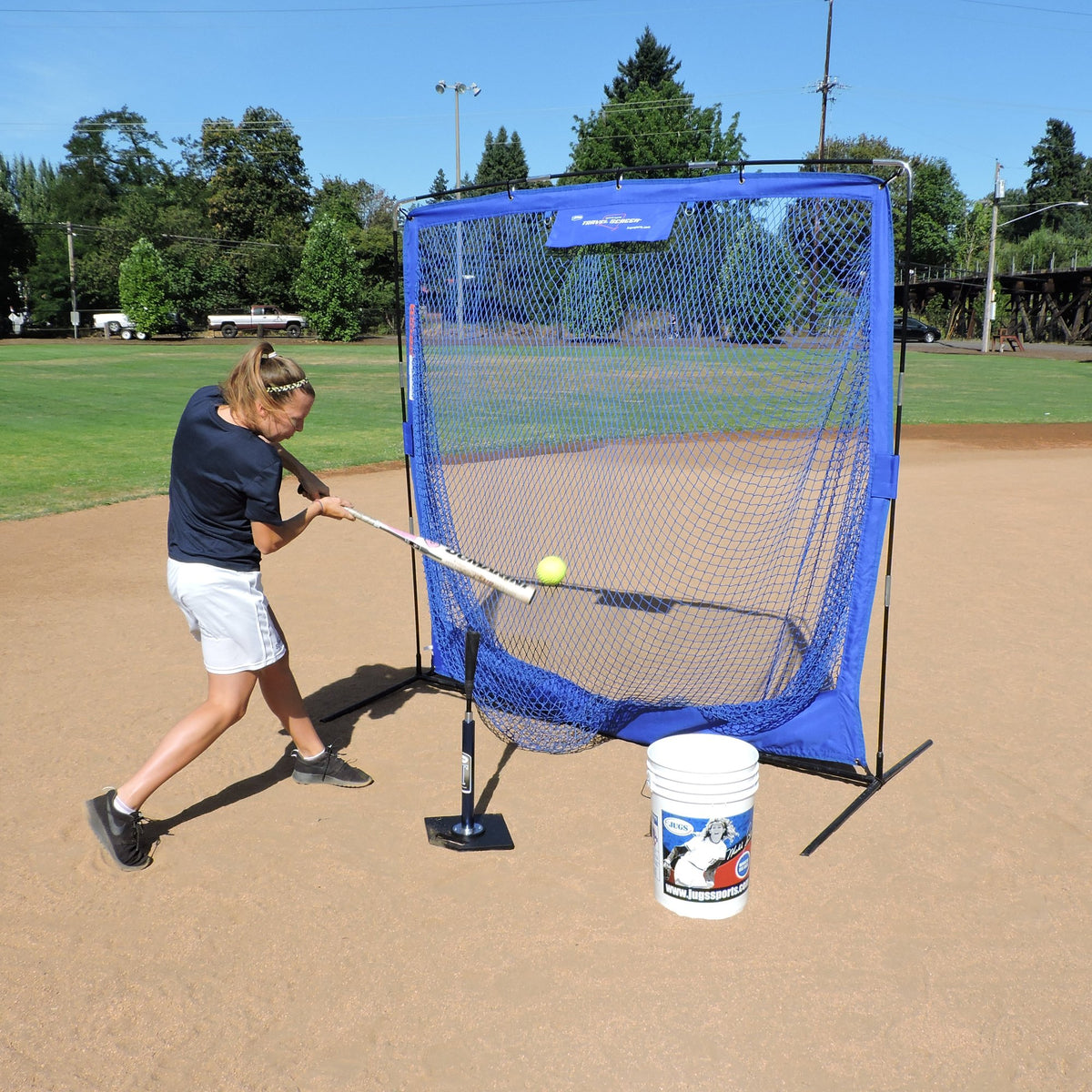 A softball player hitting a ball off a tee into the JUGS Complete Practice Travel Screen on an infield.