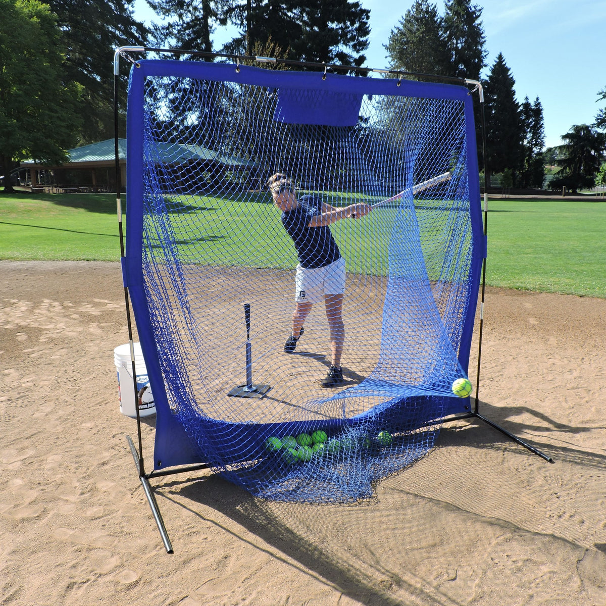 A softball player swinging at a ball off a tee in front of the JUGS Complete Practice Travel Screen.