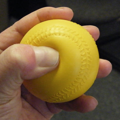 A hand squeezing a yellow foam baseball with molded seams, showing how soft it is, against a dark background.
