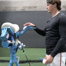 Adley Rutschman feeding a baseball into the JUGS mini3 Baseball Pitching Machine inside an indoor batting cage.