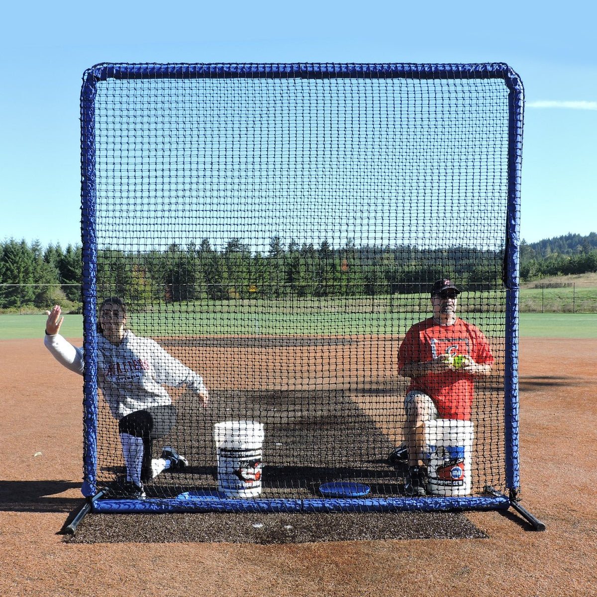 Two coaches seated behind the JUGS Protector Blue Series 8-Foot Fungo Screen on a dirt infield.
