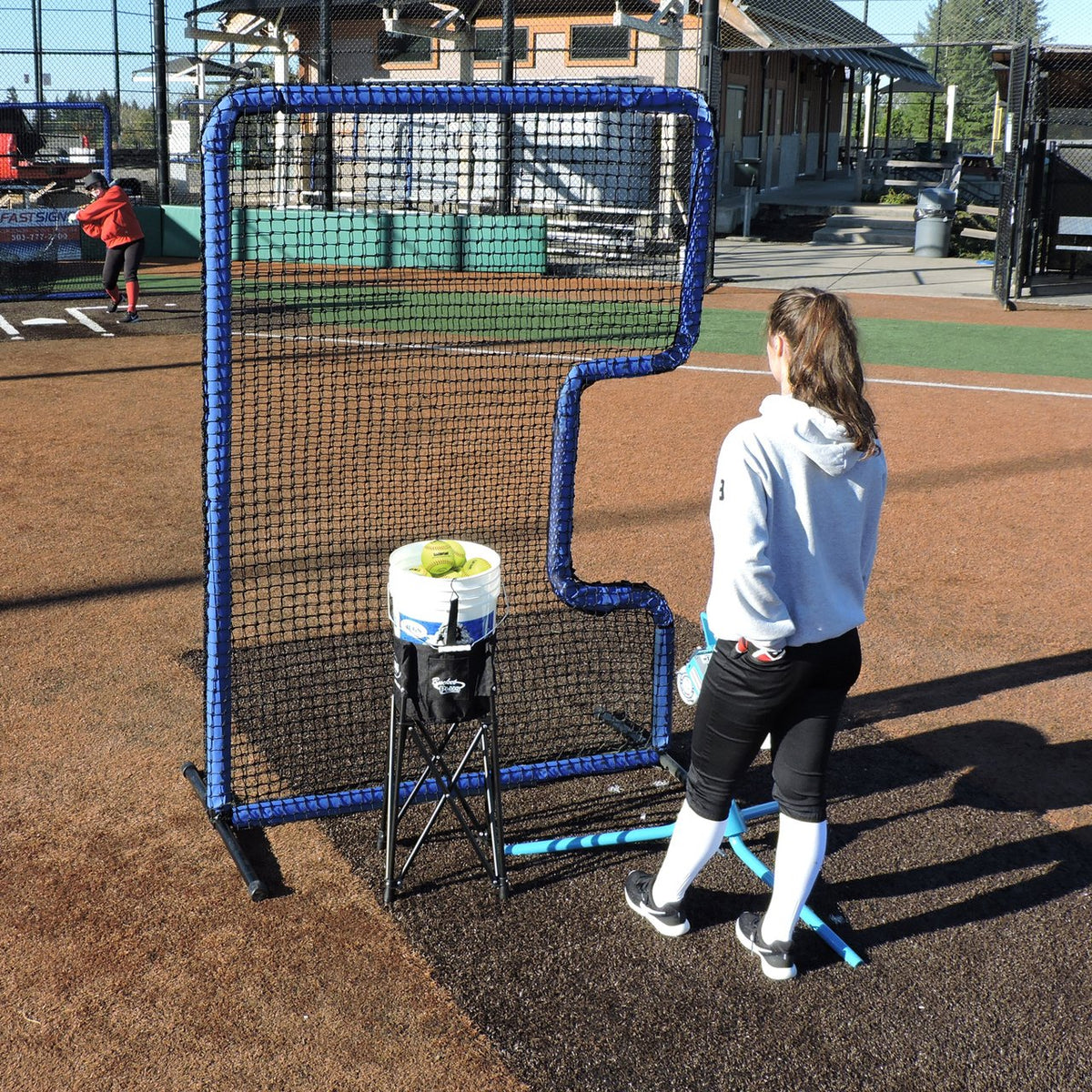A softball player standing behind the JUGS Protector Blue Series C-Shaped Softball Screen with a ball bucket on a dirt infield.
