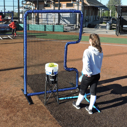 A softball player standing behind the JUGS Protector Blue Series C-Shaped Softball Screen with a ball bucket on a dirt infield.