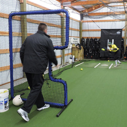 A coach tossing to a softball hitter from behind the JUGS Protector Blue Series C-Shaped Softball Screen in an indoor cage.
