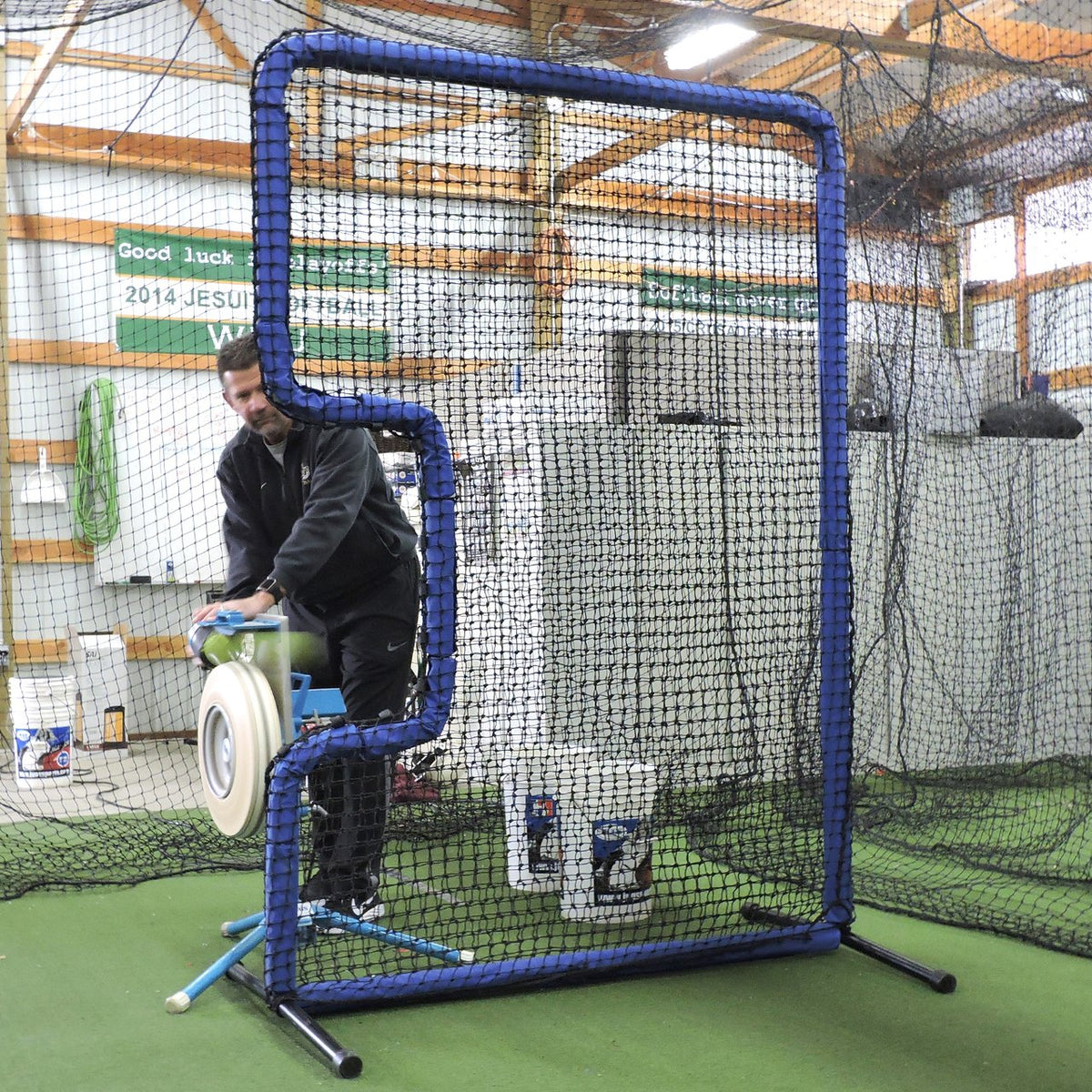 A coach feeding a JUGS pitching machine behind the JUGS Protector Blue Series C-Shaped Softball Screen indoors.