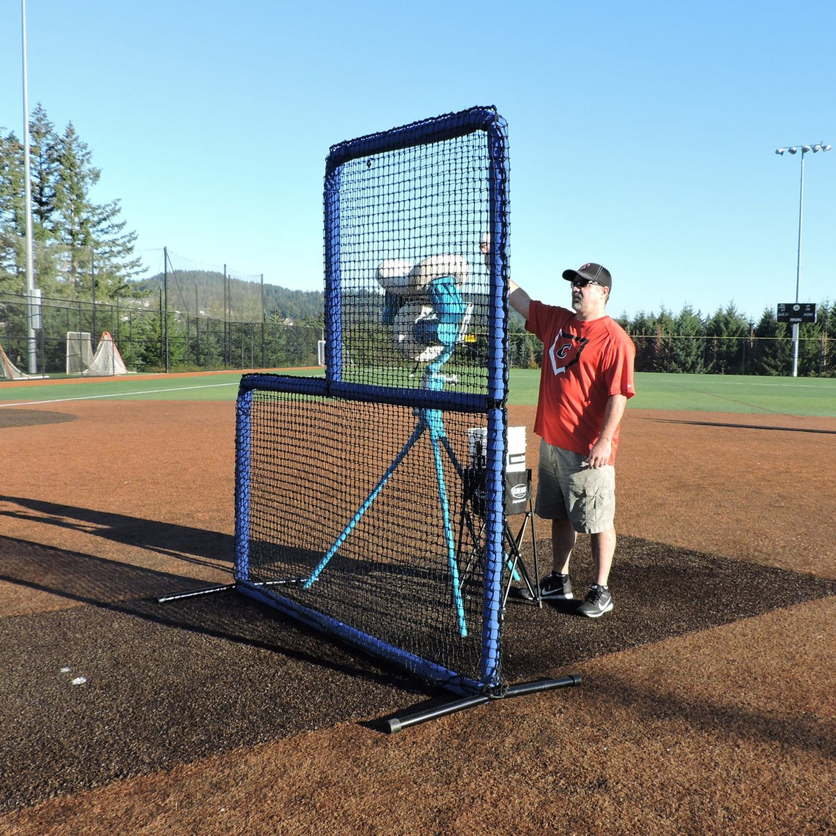 A coach feeding a pitching machine behind the JUGS Protector Blue Series L-Shaped Pitchers Screen on a baseball mound.