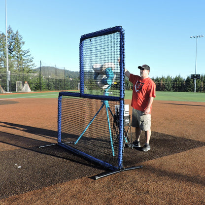 A coach feeding a pitching machine behind the JUGS Protector Blue Series L-Shaped Pitchers Screen on a baseball mound.