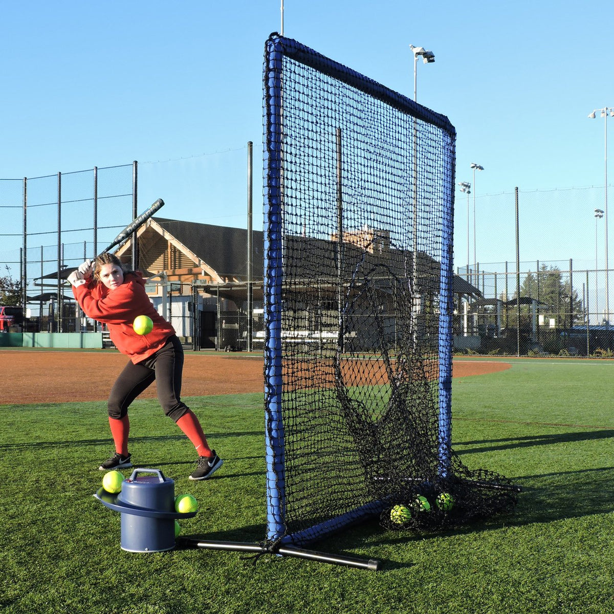 A softball player hitting into the JUGS Protector Blue Series Square Screen with Sock-Net next to a ball bucket.