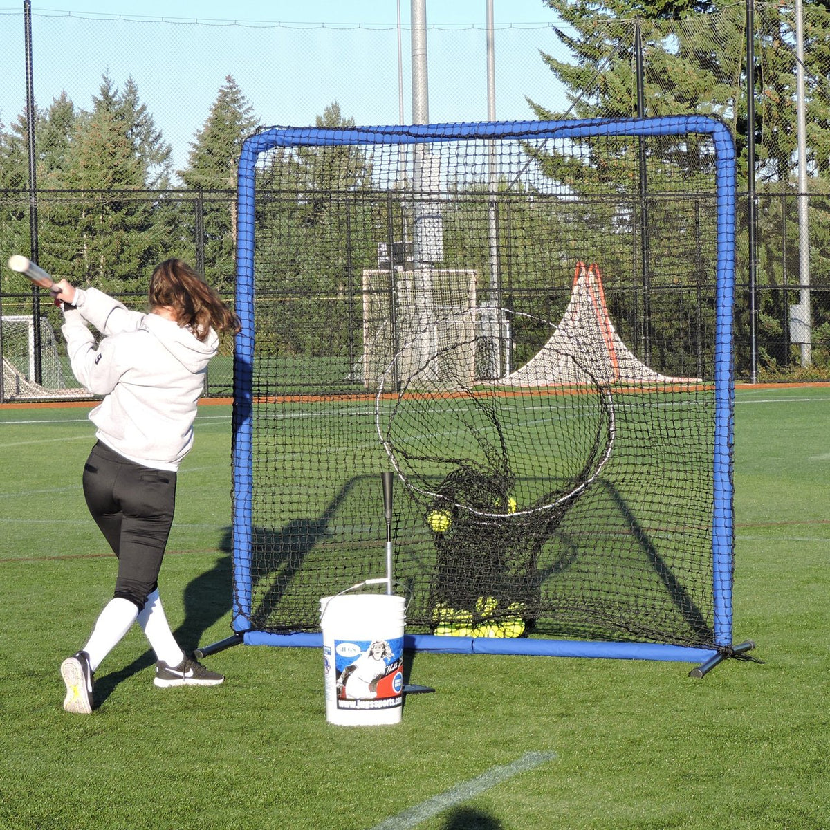 A softball player swinging at the JUGS Protector Blue Series Square Screen with Sock-Net on a turf field.
