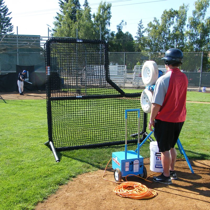 A player in a batting helmet feeding a pitching machine behind the JUGS Protector Series L-Shaped Screen.